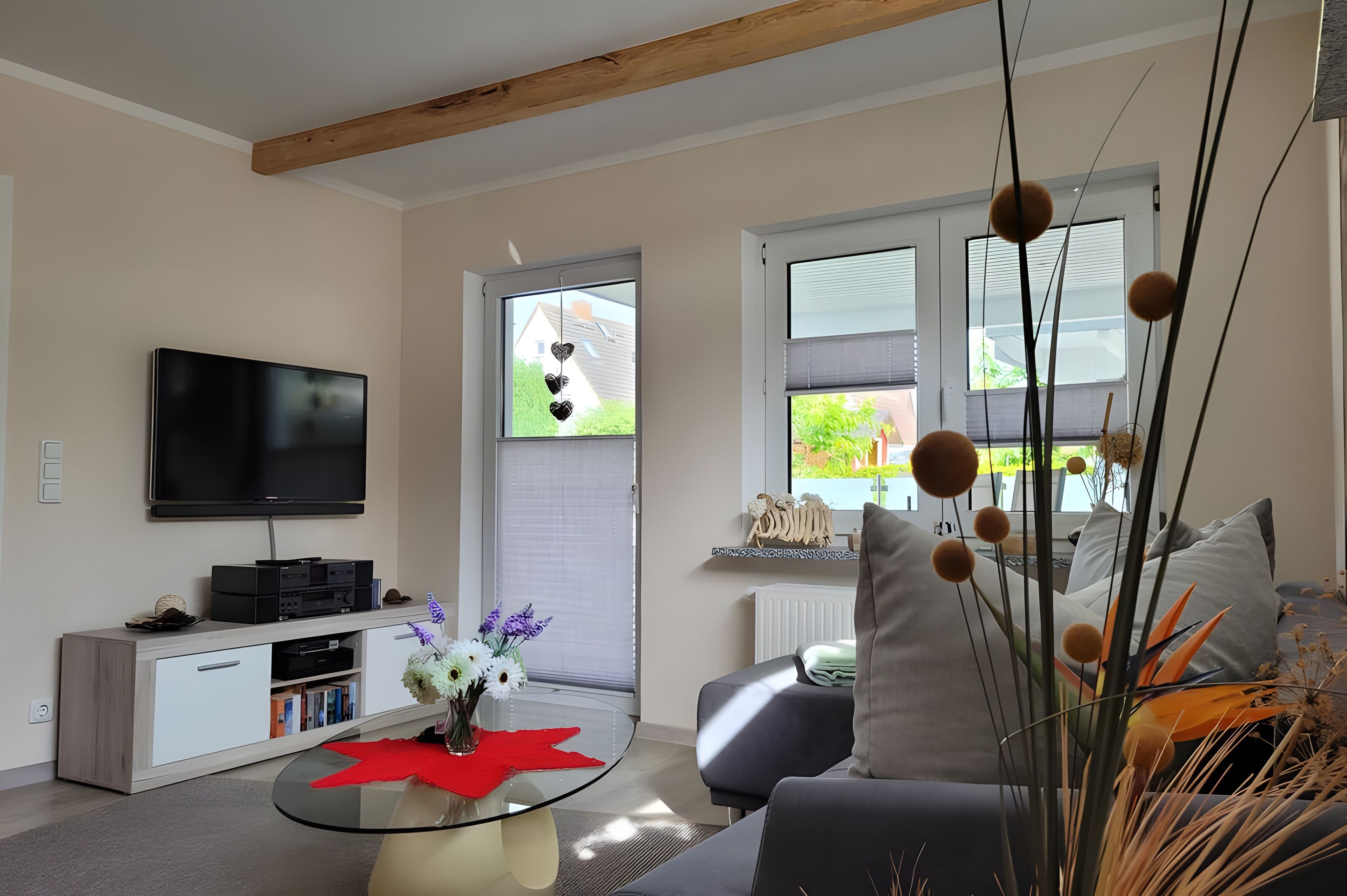 Ein modernes Wohnzimmer mit einem Flachbildfernseher, einem Glastisch mit einer roten Unterlage und einer Couch mit dekorativen Kissen.