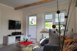 A modern living room with a flat-screen TV, a glass coffee table with a red placemat, and a sofa with decorative pillows. - Apartment 'Ferienwohnung Zeitlos' (Stralsund)