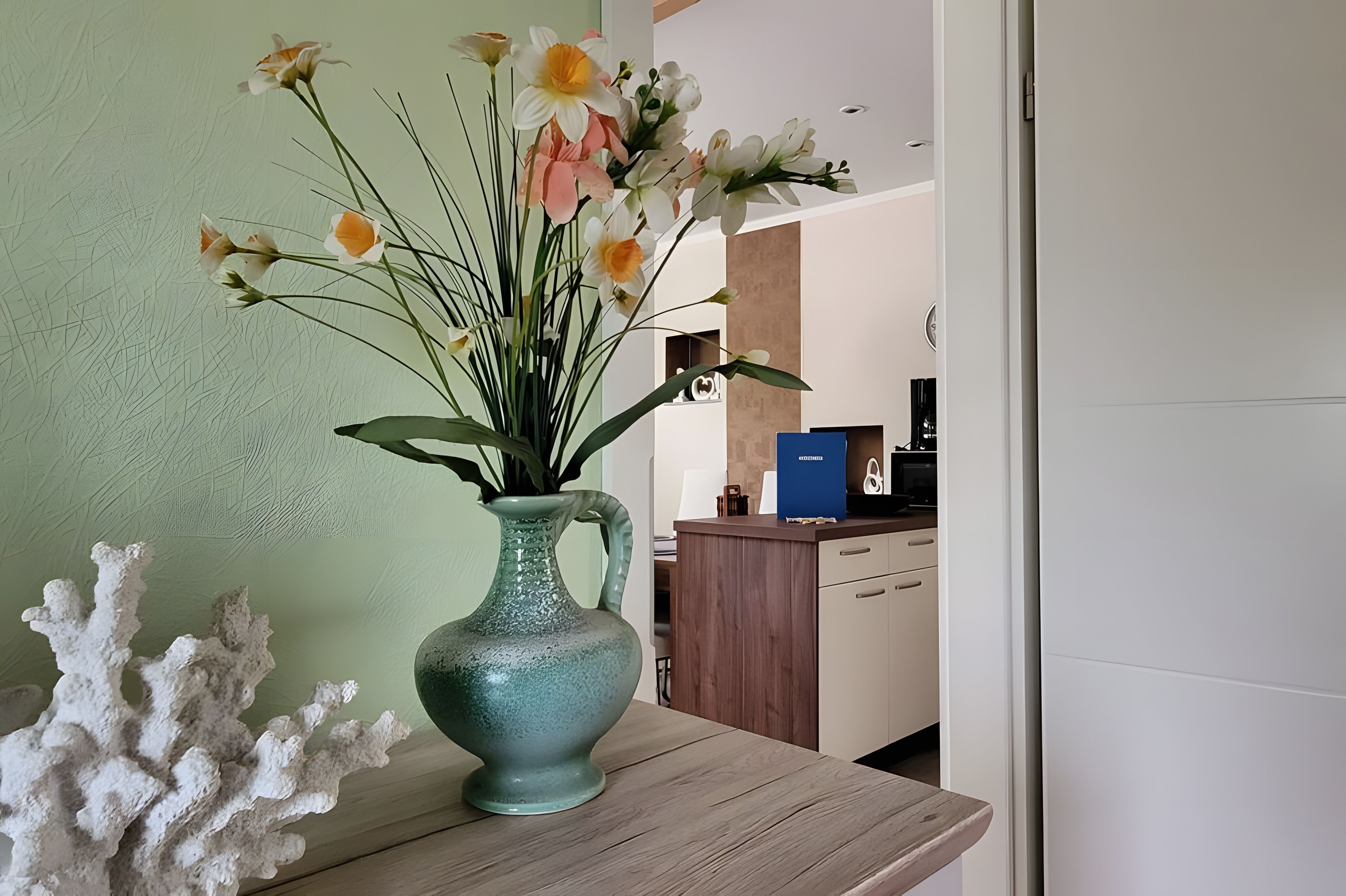 Un vaso con fiori su un tavolo di legno in una stanza con una parete verde e un'area cucina sullo sfondo.