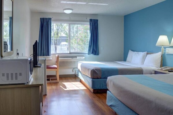 A hotel room with a bed, a desk with a computer, a chair, a window with curtains, and a lamp.