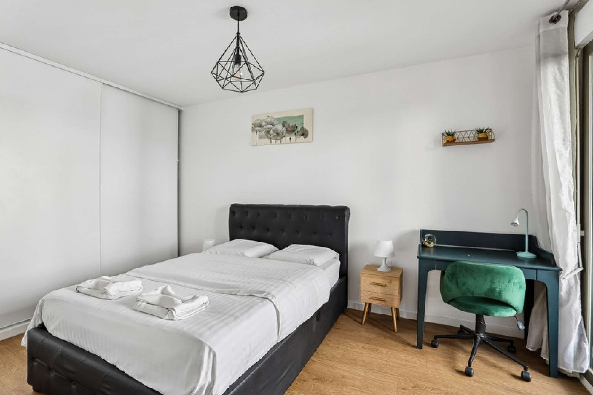 A bedroom with a bed, bedside table, desk, chair, and a hanging light fixture.