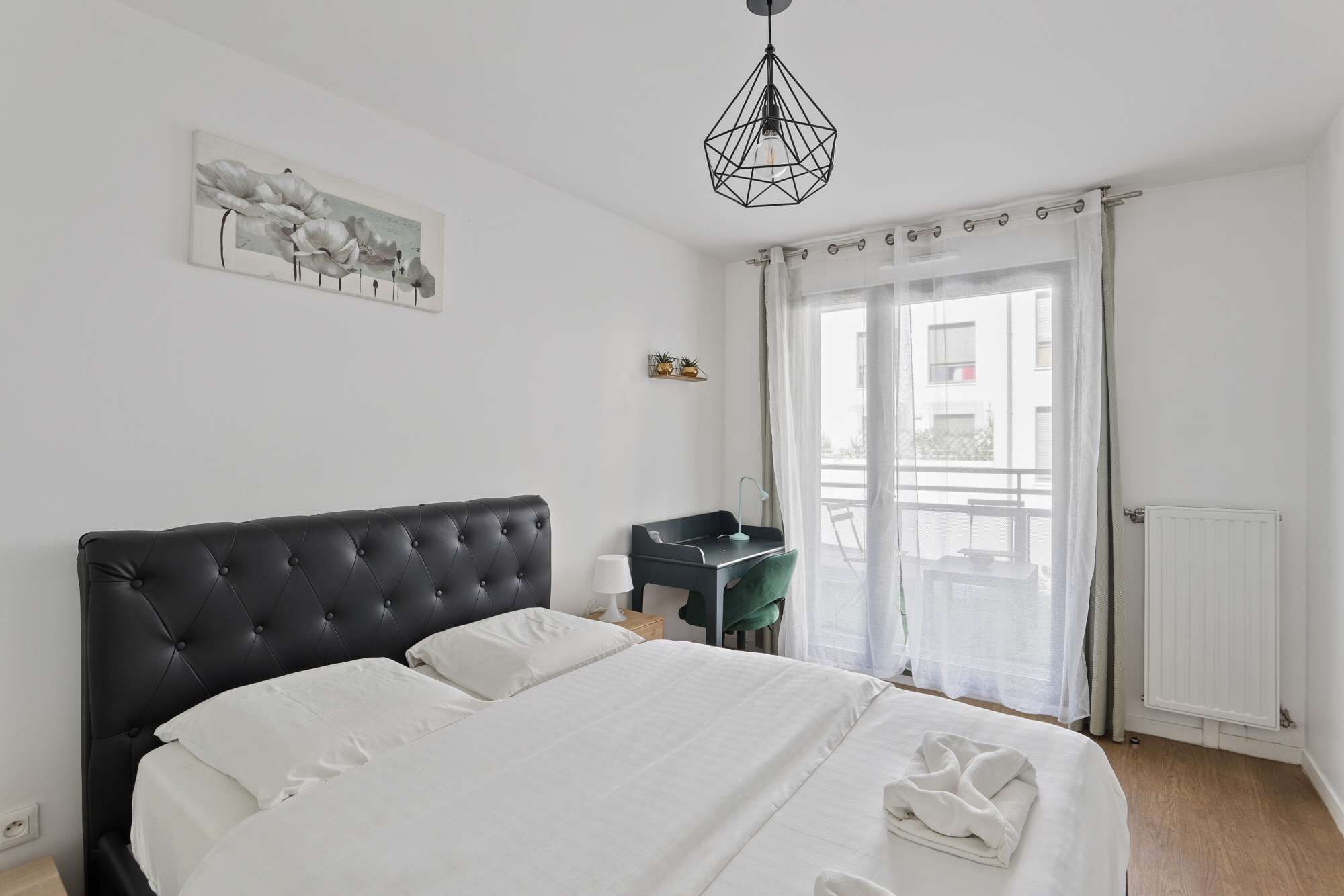 Een slaapkamer met een groot bed, een bureau, een stoel, een hanglamp en een raam met gordijnen.