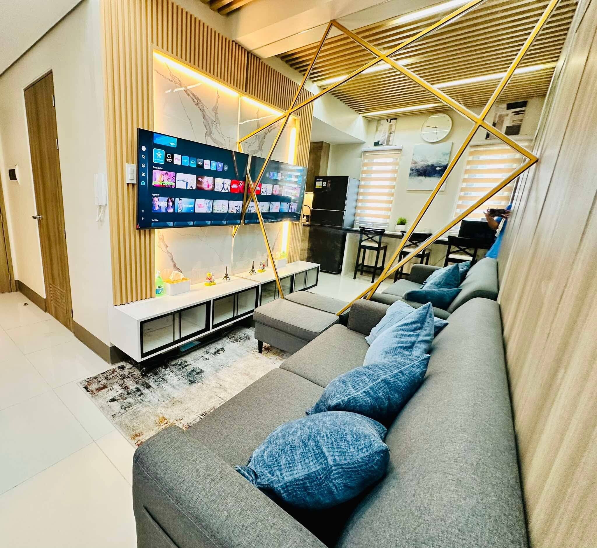 Ein modernes Wohnzimmer mit einem großen Flachbildfernseher an der Wand, einem grauen Sofa mit blauen Kissen und einer Holzdachverkleidung mit Latten.