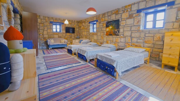 A room with a stone wall, two beds with blue bedding, a sofa, a wooden dresser, and a television mounted on the wall. - Pietra & Mare – Authentic Stone Villa Sea & Nature (Milas)