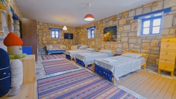 A room with a stone wall, two beds with blue bedding, a sofa, a wooden dresser, and a television mounted on the wall.