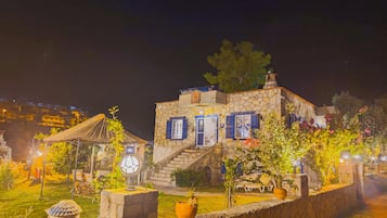 Een gebouw van steen met blauwe luiken, een rieten dakconstructie en een tuin met potplanten en een vogelhuisje.