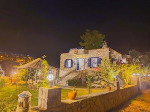 A stone building with blue shutters, a thatched roof structure, and a garden with potted plants and a birdhouse.