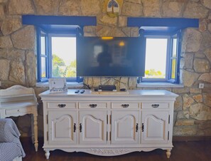A room with a stone wall, a large flat-screen TV mounted above a white cabinet with multiple drawers, and two windows with blue frames.