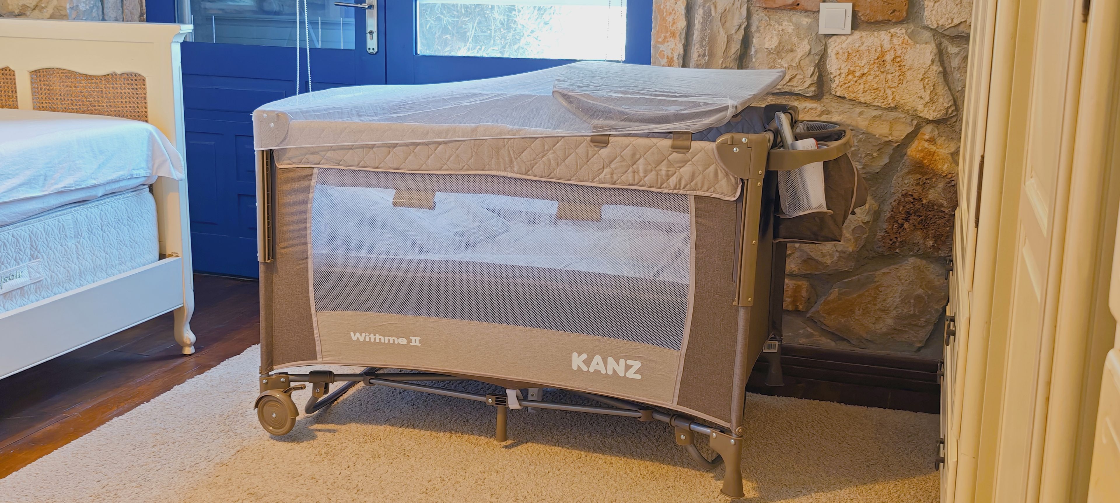 A baby crib with a canopy labeled 'Withme II' and 'KANZ' in a room with a stone wall and a blue door.