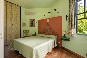 A bedroom with a bed, a nightstand, a wall-mounted air conditioner, a window with a cross, and a door with patterned curtains. - Villa "Villino Arsella" with mountain view, balcony, and air conditioning (Pietrasanta)