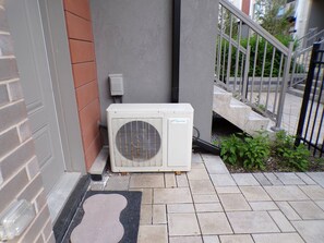 En airconditionenhed monteret på en bygningsvæg, en dørmåtte og en trappe med gelænder.
