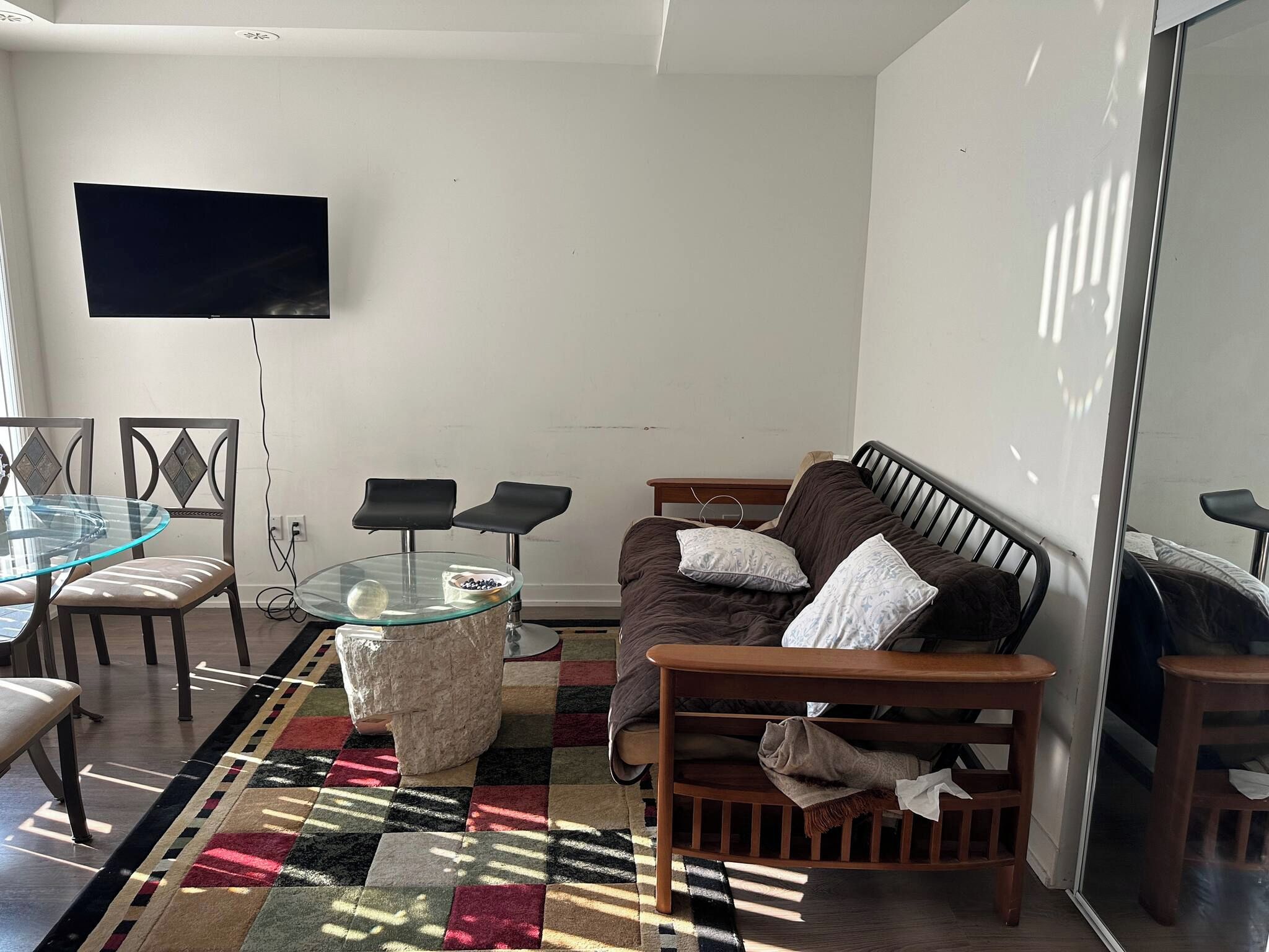A living room with a glass-top coffee table, a sofa with cushions, a TV mounted on the wall, and a patterned rug.