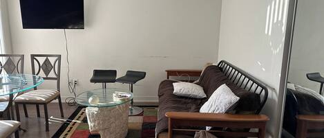 A living room with a glass-top coffee table, a sofa with cushions, a TV mounted on the wall, and a patterned rug.