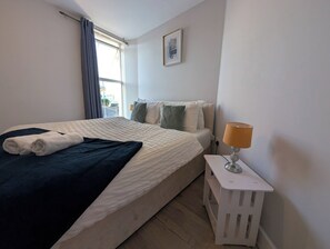 A hotel room with a bed, a bedside table with a lamp, a window with curtains, and a painting on the wall. - Harbour Haven Dover Road Folkestone (Kent)