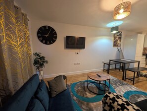 Ein Wohnzimmer mit einer Couch, einem Fernseher, einer Pflanze und einem Essbereich inklusive Tisch und Stühlen.
