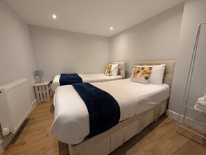 Ein Hotelzimmer mit zwei Betten, einem Heizkörper, einem kleinen Tisch mit einer Lampe und einem Regal mit Handtüchern.