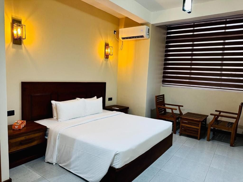 Habitación de hotel con una cama grande, mesitas de noche, un escritorio, dos sillas, un equipo de aire acondicionado y una ventana con persianas.