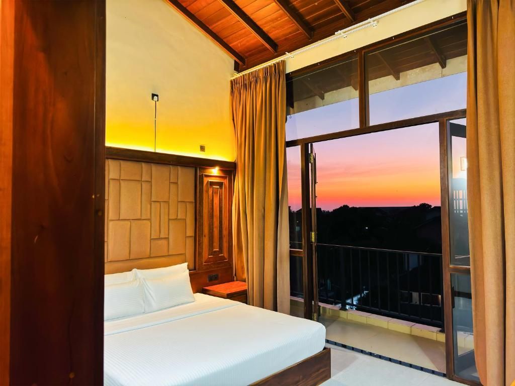 A hotel room with a large bed, a nightstand, and a view of the sunset through a balcony.