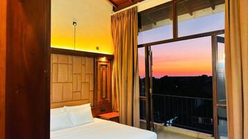 A hotel room with a large bed, a nightstand, and a view of the sunset through a balcony.