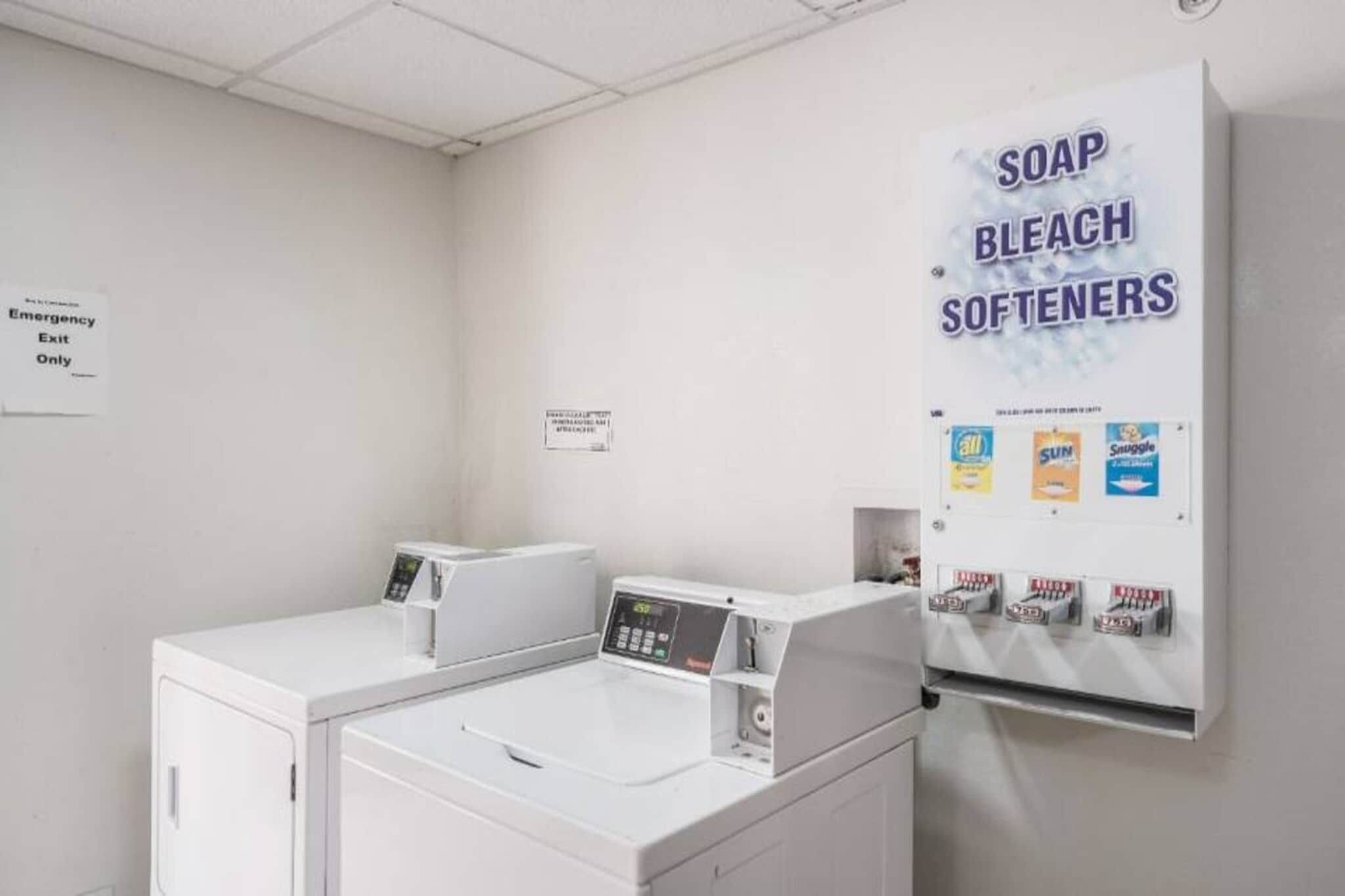 Een wasserette met wasmachines en drogers, een bordje voor de nooduitgang, en een wanddispenser voor zeep, bleekmiddel en wasverzachter.
