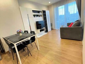 A modern living space with a dining area, a sofa, a TV, and a window with curtains. - Luxurious 2-Bedroom Apartment Near Chao Phraya River (Bangkok)