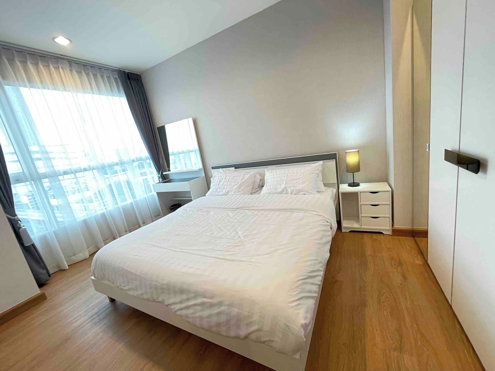 A modern hotel room with a large bed, a bedside table with a lamp, and a view of the city through the window.