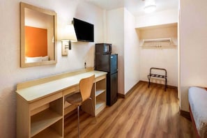 A compact room with a wooden desk, a chair, a black refrigerator, a television mounted on the wall, and a bed with a metal frame.