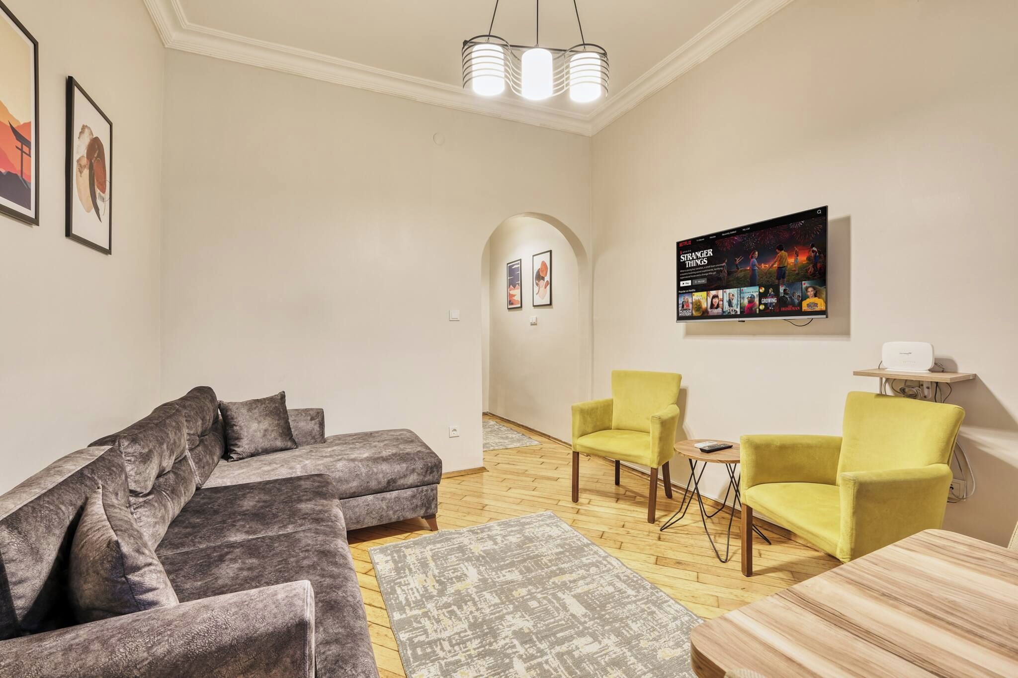 Een moderne woonkamer met een grijze modulaire bank, twee gele fauteuils, een houten salontafel en een flatscreen televisie aan de wand.