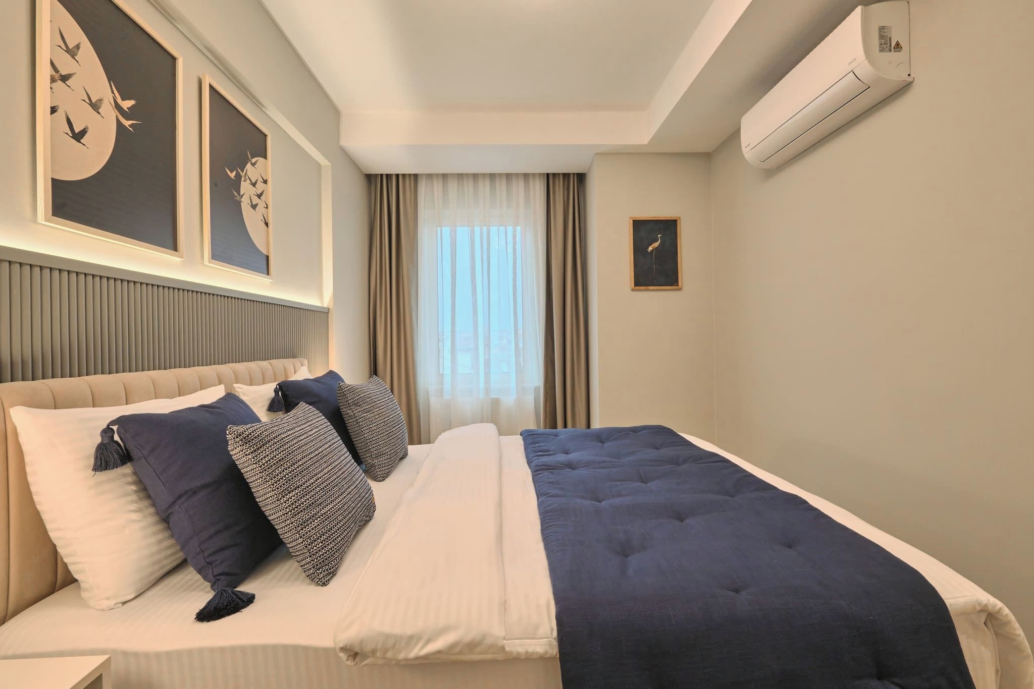 A hotel room with a bed, a headboard, a window with curtains, and wall-mounted air conditioning.