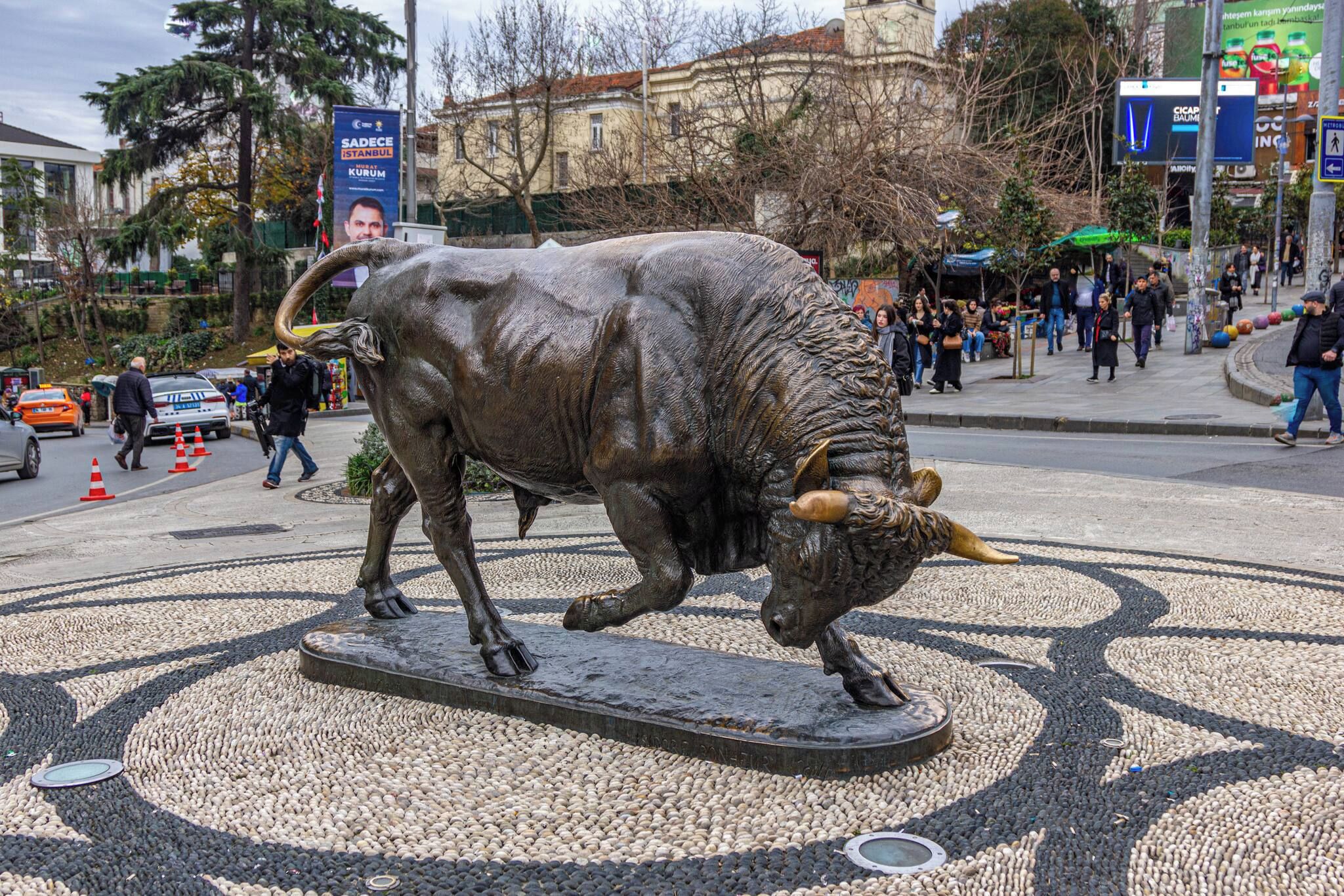 Une statue de taureau se trouve sur une place publique, entourée d’un pavage aux motifs géométriques.