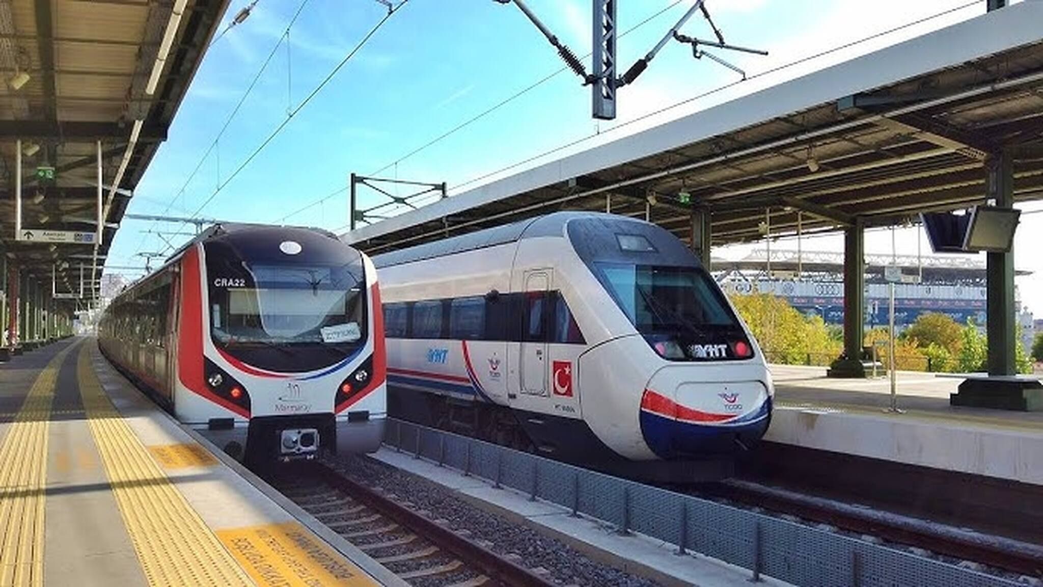 Deux trains sur le quai d’une gare, avec des lignes électriques aériennes et un ciel dégagé.