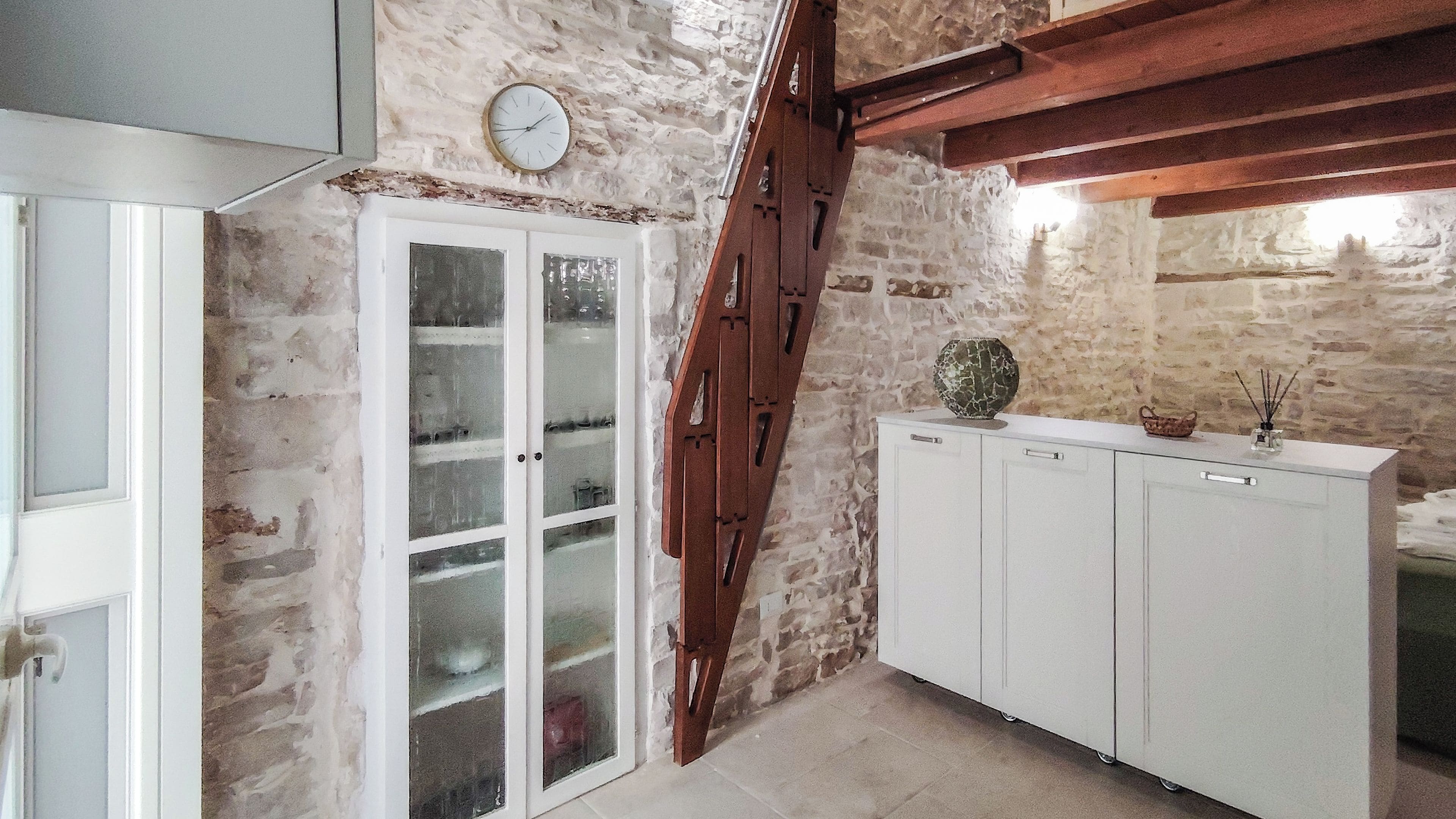 Una habitación con pared de piedra, escalera de madera, un armario blanco y una puerta con paneles de vidrio esmerilado.