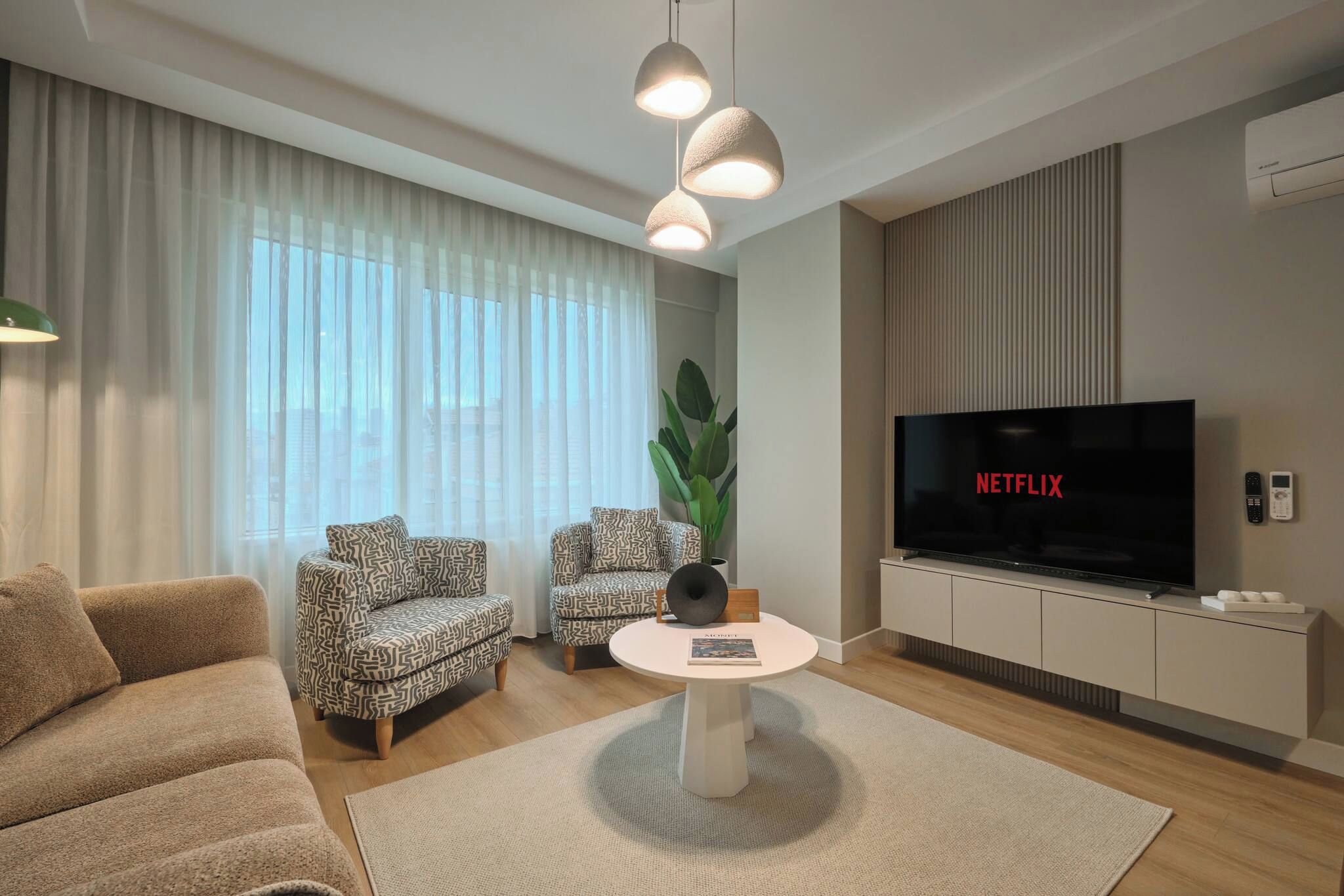 Una sala de estar moderna con un sofá, dos sillones estampados, una mesa de centro, un televisor y una planta grande.