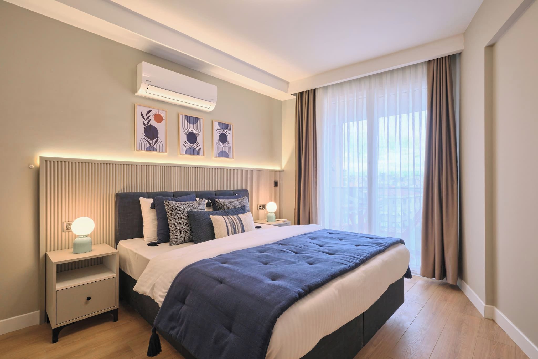 Habitación de hotel moderna con una cama grande, lámparas de noche, aire acondicionado montado en la pared y vista a la ciudad por la ventana.