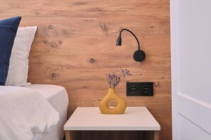 Un dormitori amb un capçal de fusta, una làmpada de nit, un vas amb flors seques i una presa de corrent a la paret.