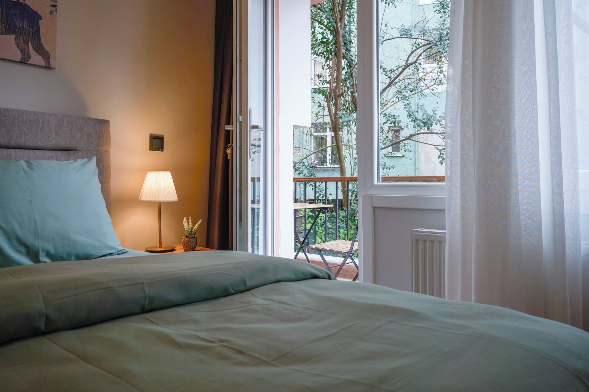 A bedroom with a bed, a bedside lamp, a window with a view of greenery, and a door leading outside.