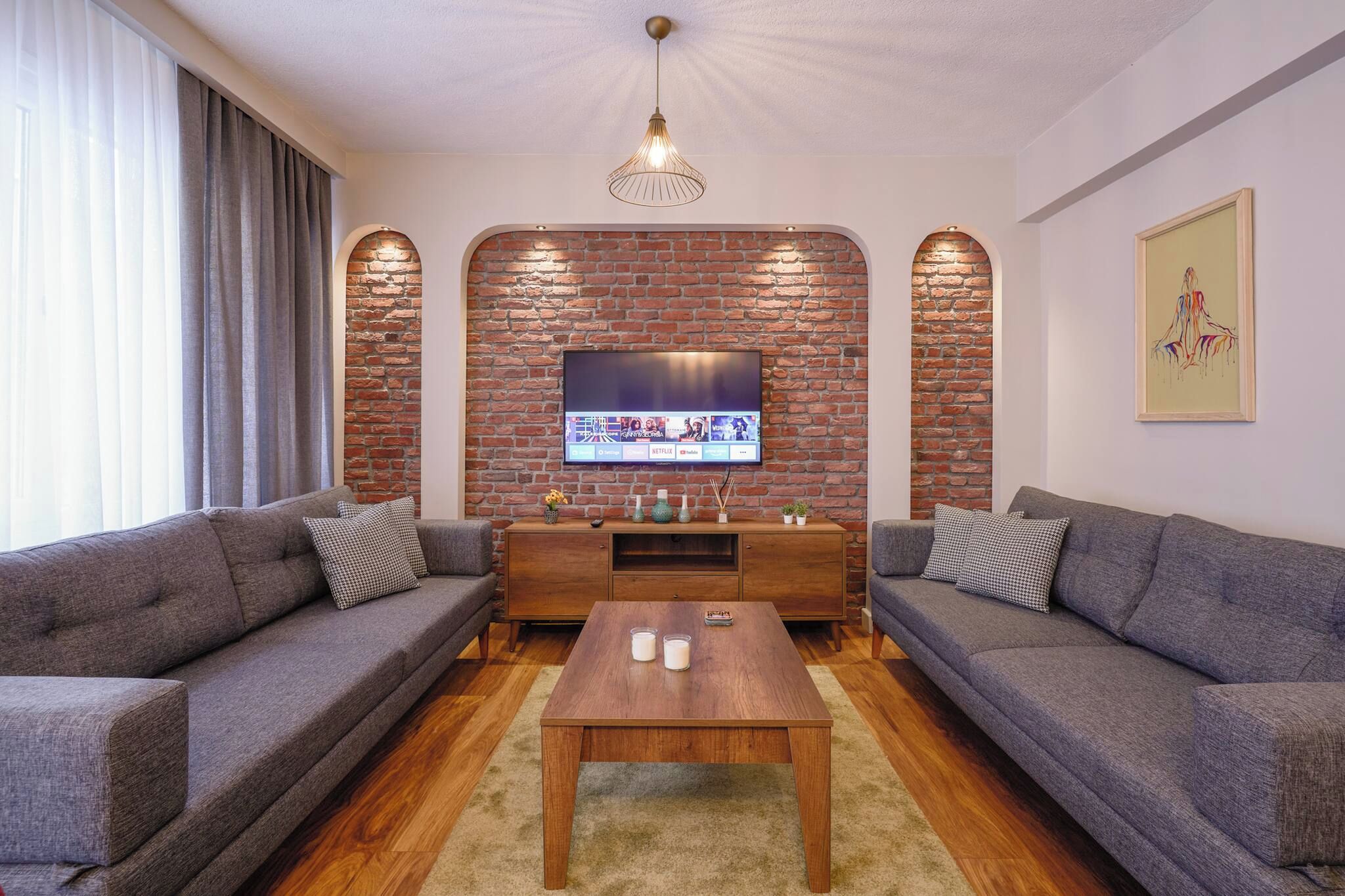 A living room with two sofas, a coffee table, a TV mounted on a brick wall, and a chandelier.