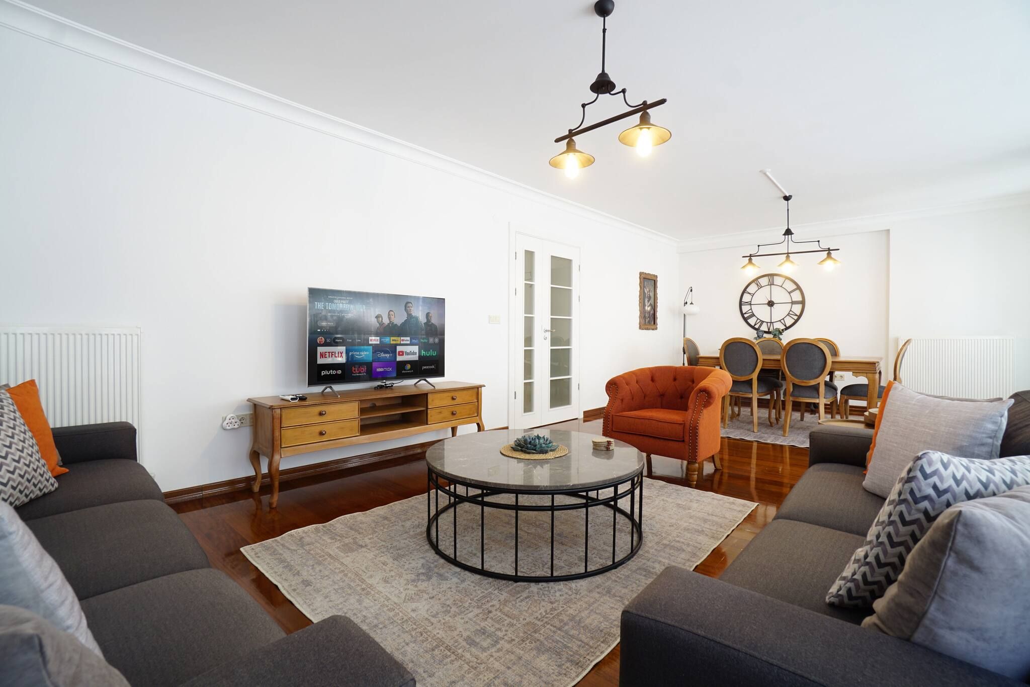 Ein modernes Wohnzimmer mit Flachbildfernseher, rundem Couchtisch, Sofa, Sessel und einem Essbereich mit Holztisch und Stühlen.