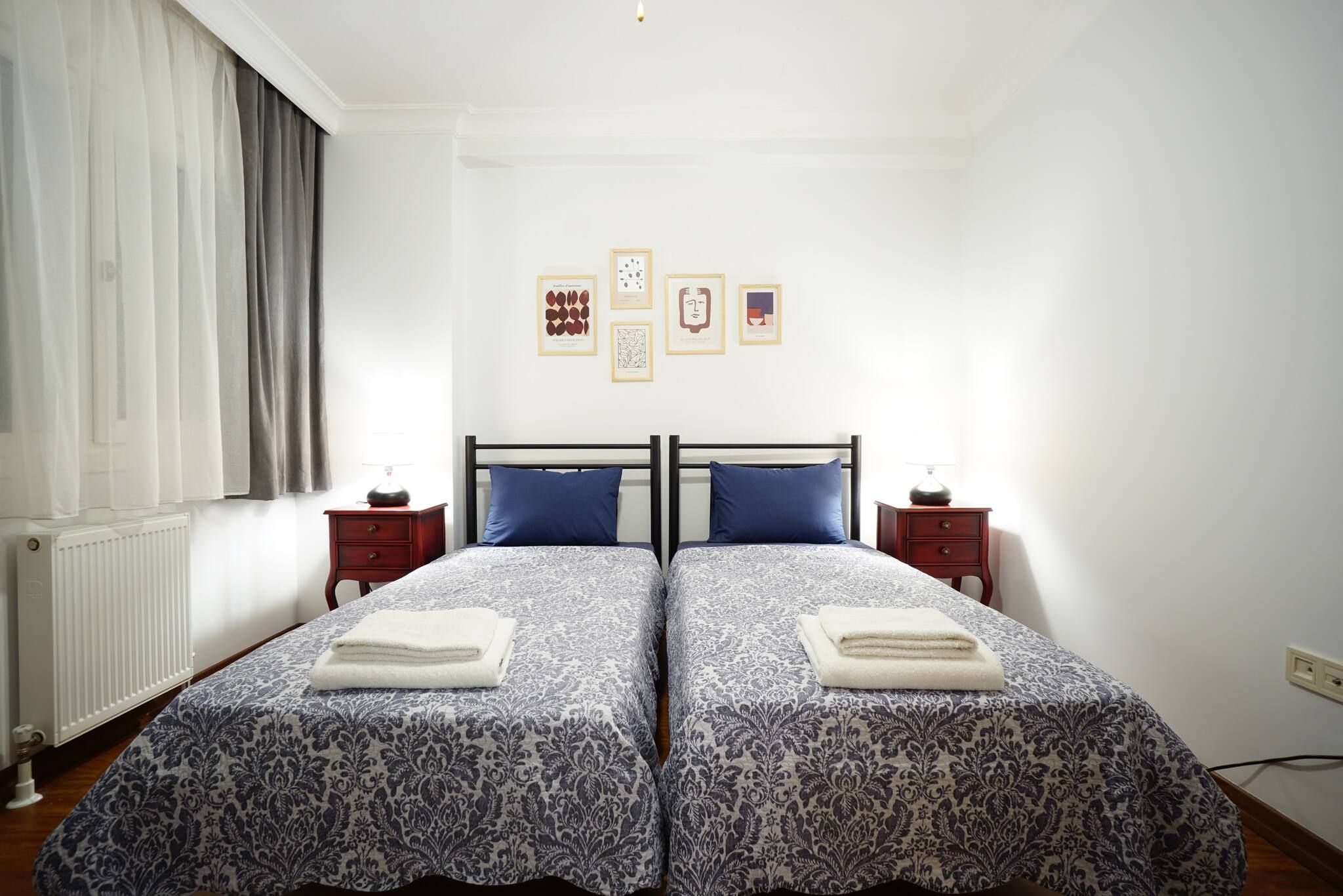 A double bed with a patterned bedspread, two bedside tables, a radiator, and a window with curtains.