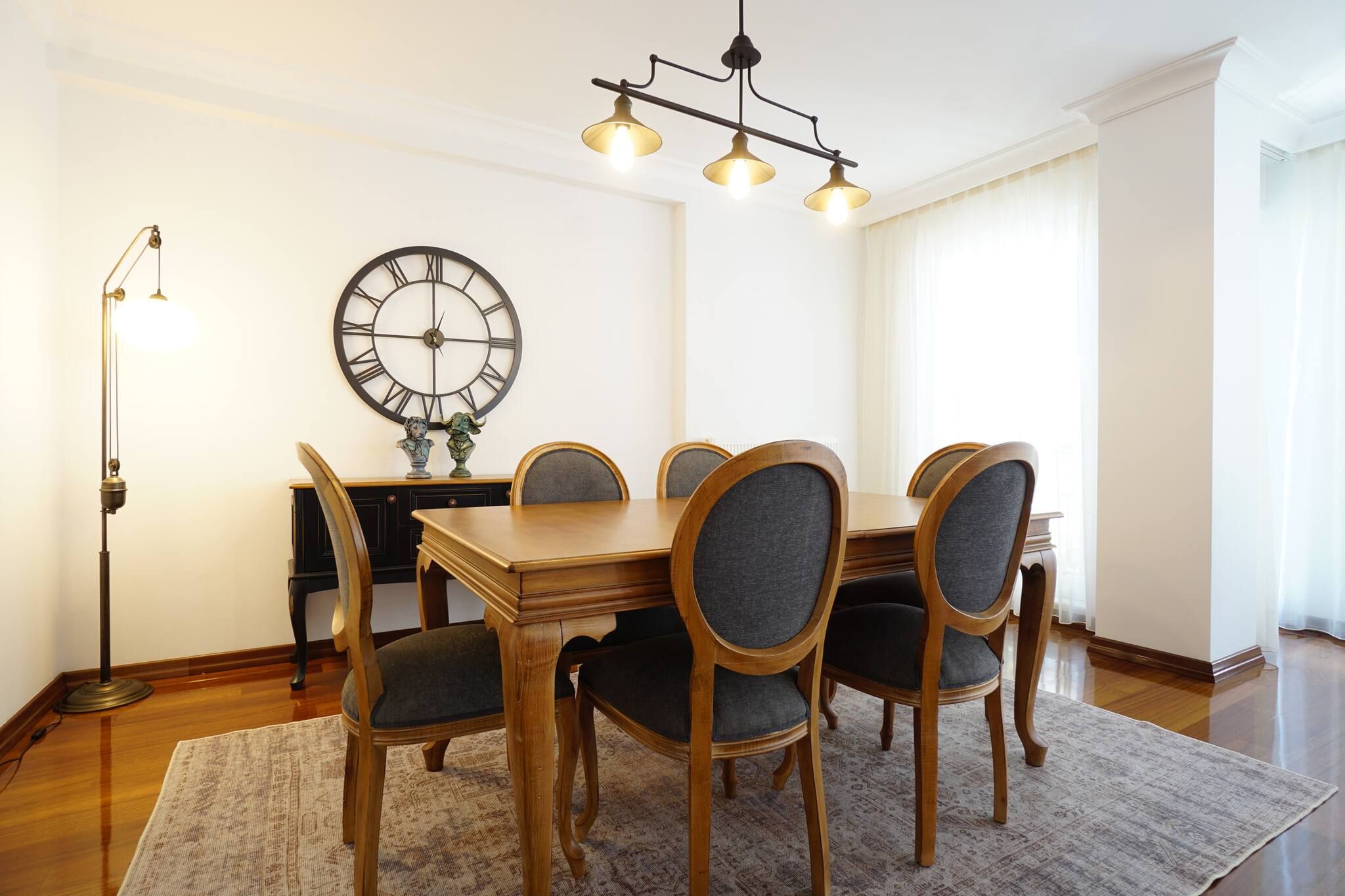 A dining room with a wooden table and chairs, a large wall clock, and a floor lamp.