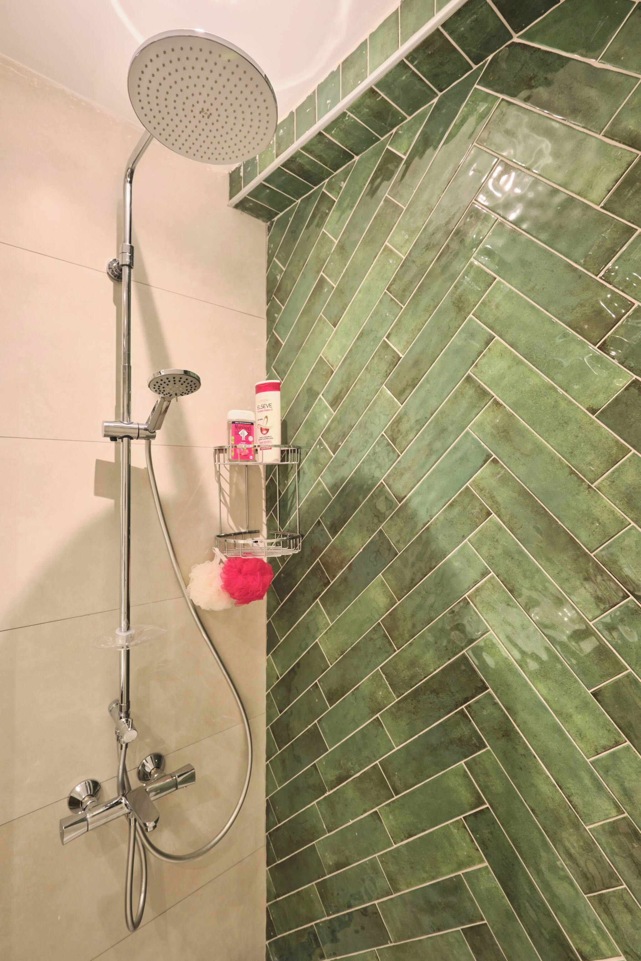 Une douche équipée d’un pommeau de douche à effet pluie et d’un pommeau de douche à main, un porte-serviettes avec une serviette blanche et une serviette rose, et une bouteille de shampoing et d’après-shampoing posée sur une étagère.