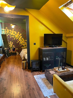 Habitació amb sòl de fusta, llit amb capçalera estampada, televisió sobre un suport, petit escalfador i paret decorativa amb dibuixos abstractes.
