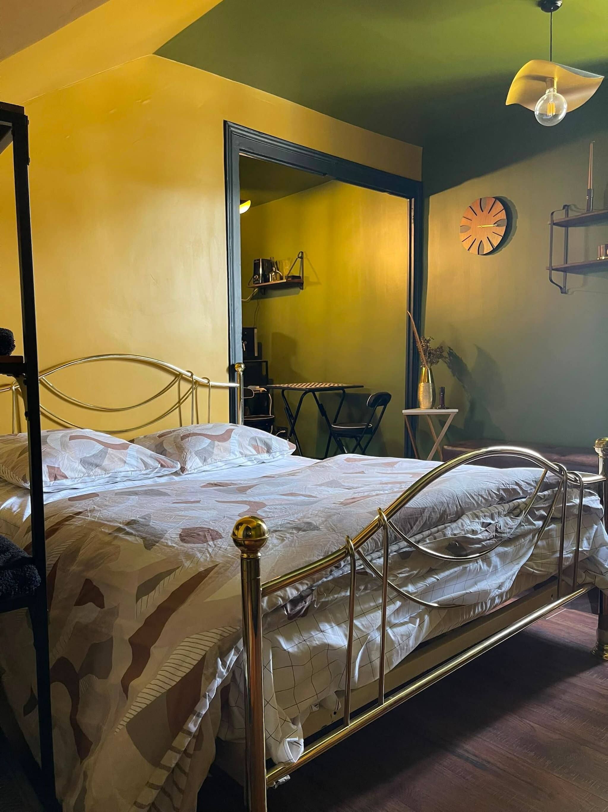 A bedroom with a four-poster bed, a small table, a chair, a clock, and a shelf with items.