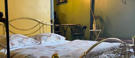 A bedroom with a four-poster bed, a small table, a chair, a clock, and a shelf with items.