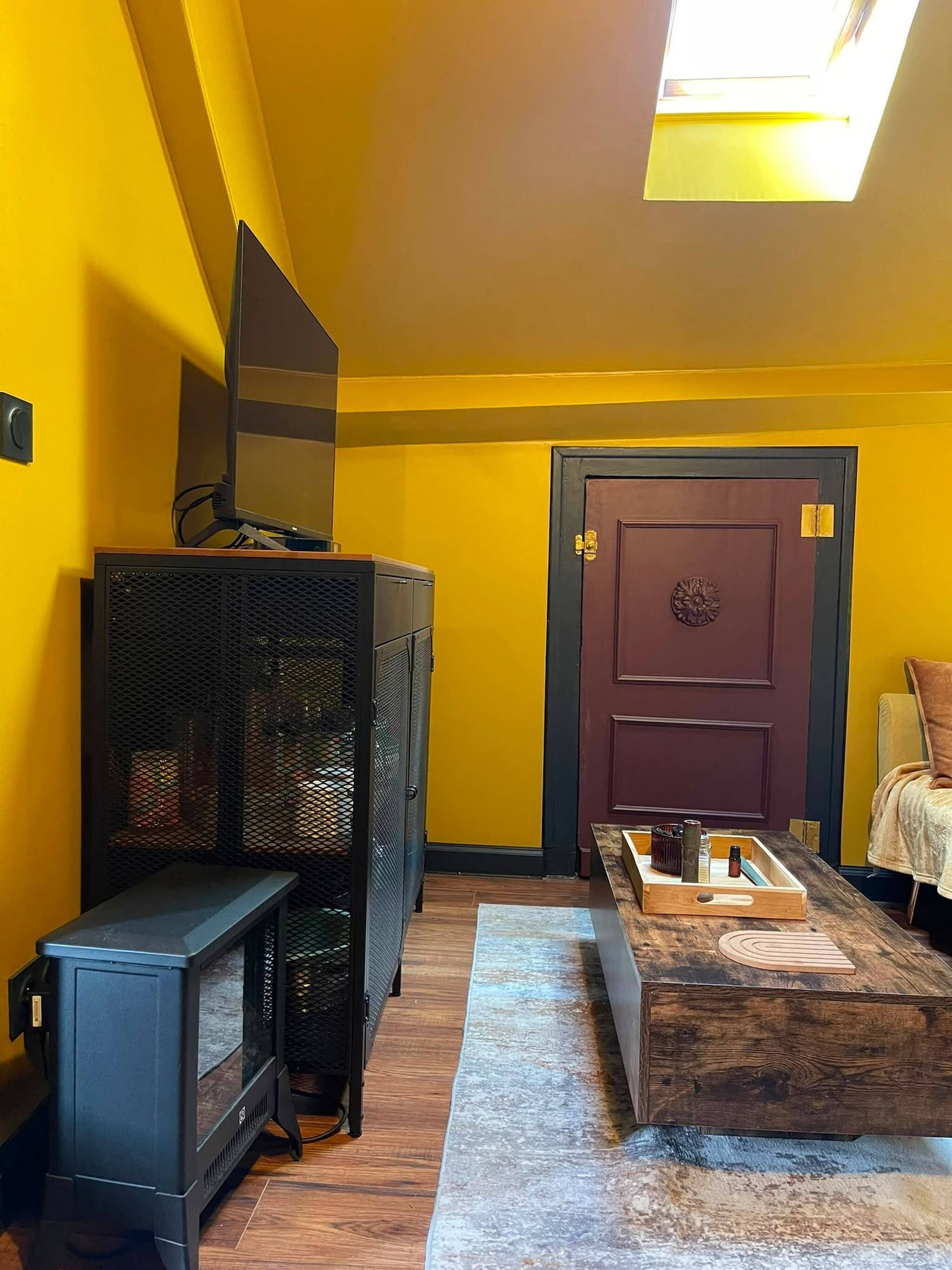 Quarto com parede amarela, porta de madeira escura, uma pequena mesa com objetos, um banco e uma televisão em um suporte.