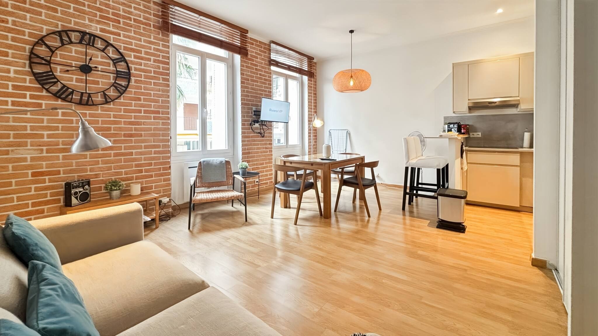 A modern living room with a brick wall, a dining area with wooden furniture, and a kitchen with light-colored cabinets.
