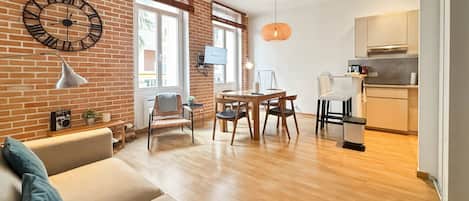 A modern living room with a brick wall, a dining area with wooden furniture, and a kitchen with light-colored cabinets.