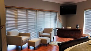 Una habitación con una cama, dos sillones, un pequeño reposapiés, una mesita de madera, un televisor montado en la pared y ventanas grandes con persianas.