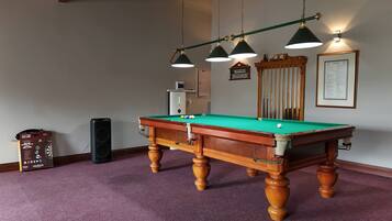 A pool table with a green felt surface, surrounded by a purple carpet. There are three hanging lamps with green shades, a wooden cabinet with a clock, and a framed menu on the wall.
