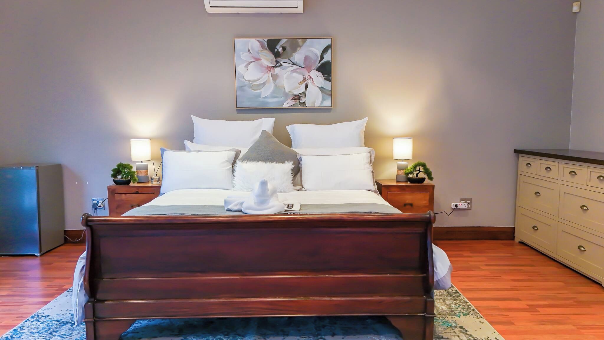 Una cama bien hecha con cabecera de madera, dos mesitas de noche con lámparas, un aire acondicionado de pared y un cuadro de flores sobre la cama.